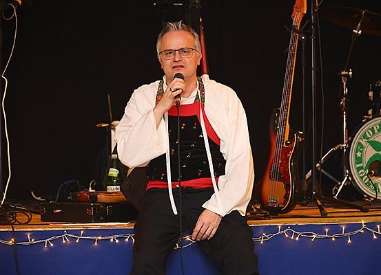 Ein Mann in einer weißen Kapuzenjacke und schwarzen Hosen sitzt auf der Bühne und hält ein Mikrofon. Hinter ihm befinden sich eine rote Gitarre, ein Schlagzeug und verschiedene Musikinstrumente.