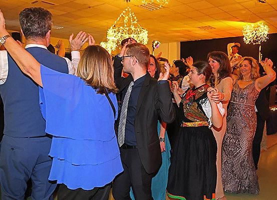 Eine Gruppe von Menschen tanzt auf einer Party, einige in formeller Kleidung. Eine Krone hängt von der Decke.