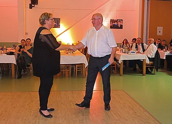 Ein Mann und eine Frau schütteln sich in einem Bankettsaal die Hände, in dem Tische und Stühle stehen. Die Gäste sitzen an den Tischen. Der Mann hält eine blaue Mappe.