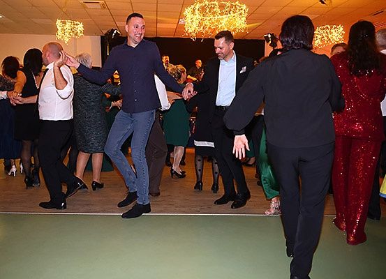 Eine Gruppe von Menschen tanzt in einem Saal mit festlicher Atmosphäre. Zwei Männer tanzen in der Mitte, während andere im Hintergrund tanzen.