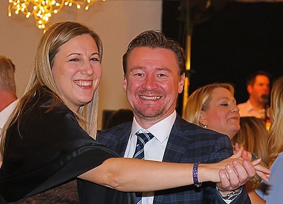 Ein lächelnder Mann und eine Frau tanzen bei einer Party zusammen, mit anderen Leuten im Hintergrund. Der Mann trägt einen Anzug und eine Krawatte, und die Frau trägt ein Armband.