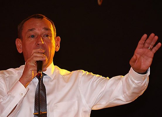 Ein Mann in weißem Hemd und Krawatte spricht in ein Mikrofon und gestikuliert mit der rechten Hand.