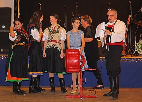 Fünf Personen in traditionellen Kostümen auf der Bühne mit einem roten Objekt, einem Mann, der ein Papier hält, und einer Frau, die mit einem Mädchen spricht. Dahinter sind ein Schlagzeug und Lautsprecher zu sehen.