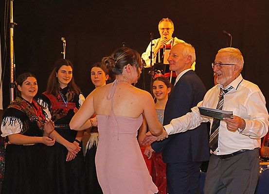 Eine Gruppe von Menschen auf einer Bühne, eine Frau in einem rosa Kleid tanzt mit einem älteren Mann. Ein Mann in Anzug und Brille hält ein Buch und ein Mikrofon. Hinter ihnen beobachten eine Gruppe von Frauen in traditionellen Kostümen.