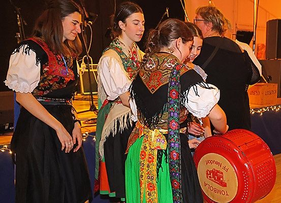 Eine Gruppe von Frauen in traditionellen Kostümen lächelt und steht auf der Bühne. Ein roter Fass steht vor ihnen. Dahinter steht ein Lautsprecher auf der Bühne.