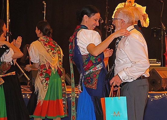 Eine Gruppe von Tänzern, einer in traditioneller Kleidung, feiert mit einem älteren Mann, der einen Geschenkbeutel hält, auf einer Bühne mit Musikern im Hintergrund.