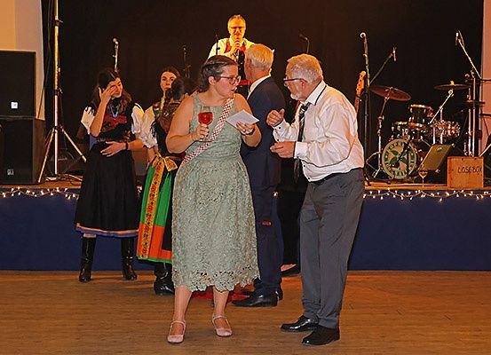 Eine Frau in einem Spitzenkleid spricht mit einem älteren Mann, der ein Glas Wein in der Hand hält, auf der Bühne. Ein Schlagzeuger und ein Sänger sind im Hintergrund.