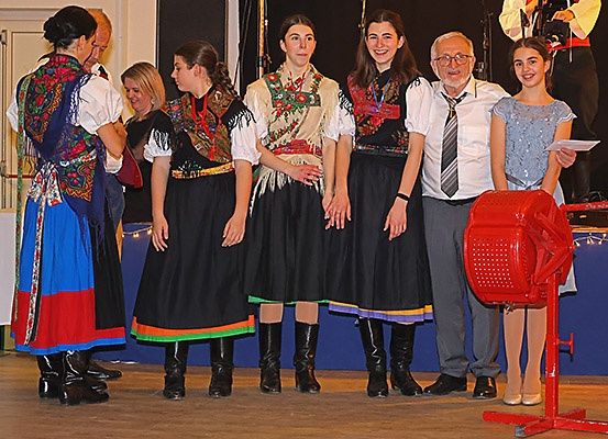Eine Gruppe von Menschen in traditioneller Kleidung steht auf einer Bühne, wobei eine Frau eine rote Maschine vor ihnen hält.