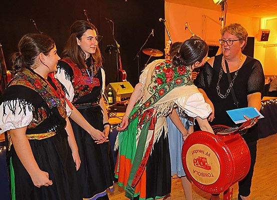 Drei Frauen in traditioneller Kleidung stehen nahe einer Frau mit Brille, die ein Papier hält, wahrscheinlich bei einer Veranstaltung mit Musikinstrumenten und einer Menschenmenge im Hintergrund.