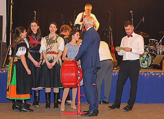 Eine Gruppe von Menschen in traditioneller Kleidung steht auf einer Bühne mit einem Mann im Anzug und einer roten Maschine. Dahinter spielt eine Band.