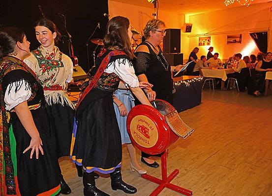 Vier Frauen in traditionellen Kostümen stehen in einem Raum, möglicherweise auf einer Party. Zwei Frauen schauen eine Frau in der Mitte an. Vor ihnen steht ein roter Ständer.