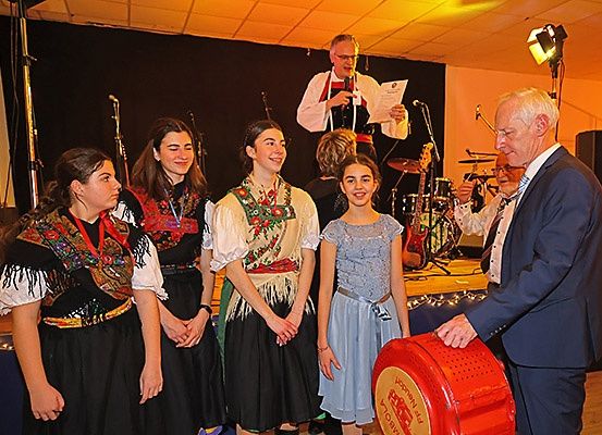 Eine Gruppe von Menschen, darunter junge Mädchen in traditioneller Kleidung, steht auf einer Bühne mit einem Mann, der ein Papier in der Hand hält. Dahinter spielt eine Band.