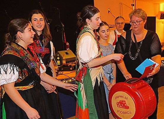 Mehrere Frauen in traditioneller Kleidung interagieren mit einer Frau, die ein Buch und einen roten Gegenstand, möglicherweise eine Spendendose, in der Hand hält. Hinter ihnen deuten ein Mikrofon und eine Gitarre auf eine Aufführung oder Veranstaltung hin.