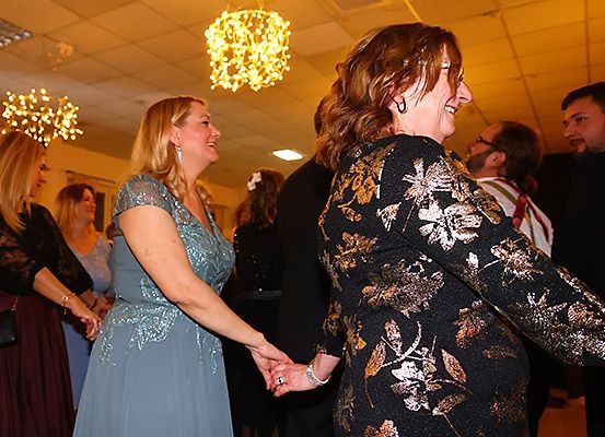Zwei Frauen, eine in einem blauen Kleid und eine andere in einem schwarzen Kleid mit Blumenmuster, halten Händchen und lachen in einem Raum mit anderen Leuten.