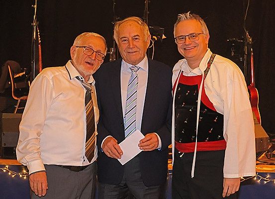 Drei Männer stehen zusammen auf einer Bühne, halten ein Papier in der Hand, mit Musikinstrumenten und einem Lautsprecher im Hintergrund.