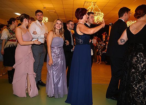 Eine Gruppe von Menschen in formeller Kleidung tanzt auf einer Tanzfläche, die von warmem Licht beleuchtet wird. Unter ihnen tanzt eine Frau in einem blauen Kleid mit einem Mann.