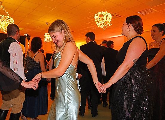 Eine Gruppe von Menschen tanzt in einem hell beleuchteten Raum mit Kronleuchtern. Eine Frau in einem silbernen Kleid lacht und hält die Hände von zwei anderen.