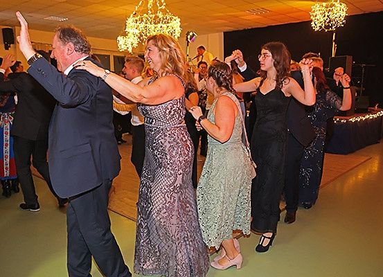 Eine Gruppe von Menschen tanzt auf einer Party. Sie sind in Abendkleidung gekleidet. Die Frau in der Mitte lächelt. Hinter ihnen steht ein Mann mit einem Mikrofon. Es gibt Kronleuchter und Lautsprecher im Bereich.