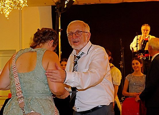 Ein älterer Mann mit Brille und Krawatte tanzt mit einer Frau in einem ärmellosen Kleid. Hinter ihnen singt ein Mann in ein Mikrofon.
