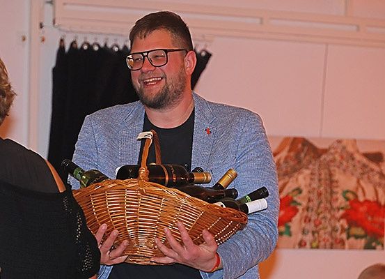 Ein Mann mit Brille und einem hellblauen Blazer hält einen Korb voller Weinflaschen und lächelt. Eine Frau mit kurzen Haaren steht hinter ihm.