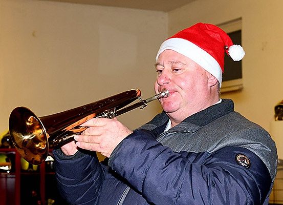 Ein Mann mit einem Weihnachtsmannhut spielt eine Posaune in einem Raum mit weißen Wänden und einem roten Regal mit Helmen.