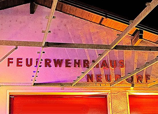 Die Außenseite eines Gebäudes, nachts beleuchtet, trägt den Schriftzug 'Feuerwehrhaus Neudorf'. Es zeichnet sich durch eine rosa Wand mit einem Metallgerüst darüber und zwei roten Türen darunter aus.