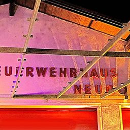 Die Außenseite eines Gebäudes, nachts beleuchtet, trägt den Schriftzug 'Feuerwehrhaus Neudorf'. Es zeichnet sich durch eine rosa Wand mit einem Metallgerüst darüber und zwei roten Türen darunter aus.