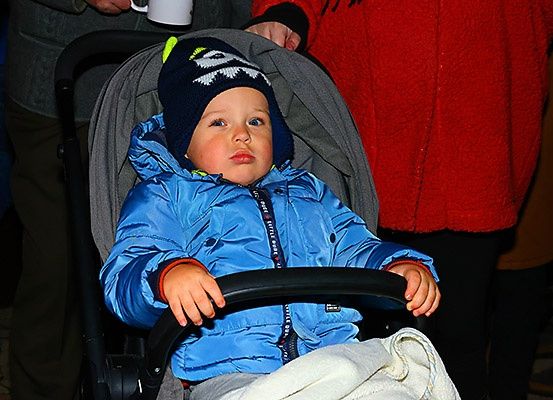 Ein kleiner Junge in einem blauen Mantel und Mütze sitzt in einem Kinderwagen mit einer weißen Decke. Hinter ihm steht eine Person in einem roten Mantel und hält eine weiße Tasse.