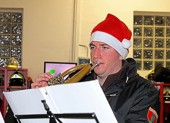 Ein Mann in einem Weihnachtshut spielt eine Posaune und schaut auf eine Musikpartitur auf einem Ständer. Im Hintergrund befinden sich Helme und Monitore.