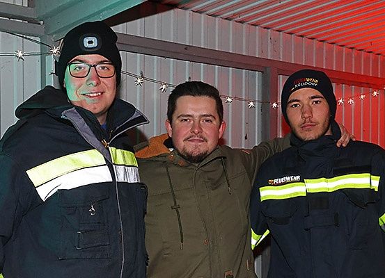 Drei Männer in Winteruniformen stehen zusammen in einem Schuppen mit roten Lichtern. Der linke Mann hat reflektierende Streifen, der mittlere trägt eine grüne Jacke und der rechte einen schwarzen Hut.