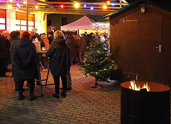 Eine Gruppe von Menschen hat sich bei Nacht um einen Weihnachtsbaum versammelt, in der Nähe eines Tisches und eines Feuerrings. Sie befinden sich wahrscheinlich auf einem Weihnachtsmarkt.