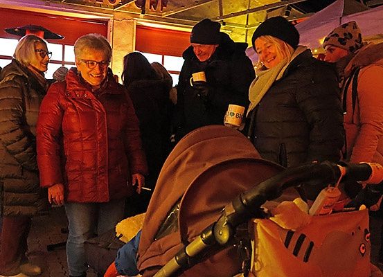 Mehrere Menschen sind drinnen versammelt und tragen warme Winterkleidung. Eine Frau hält eine Kaffeetasse, und ein Kinderwagen steht in der Nähe.
