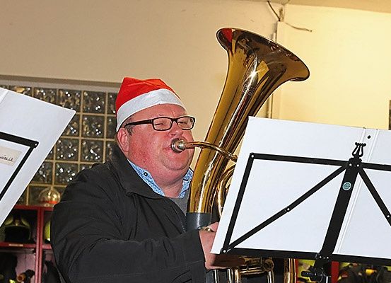 Ein Mann in einem Weihnachtsmannhut spielt Tuba und hält einen Notenständer. Der Hintergrund zeigt ein Glasfenster und Regale.