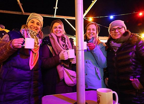 Vier Frauen in Wintermänteln und Schals lächeln für die Kamera und halten weiße Tassen. Sie stehen unter einer lila Beleuchtung, wahrscheinlich bei einer Nachtveranstaltung.