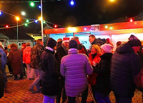 Eine Nachtszene zeigt eine Gruppe von Menschen, die sich um einen Essensstand versammelt haben, der mit bunten Lichtern geschmückt ist. Sie sind warm gekleidet, was auf eine kalte Wetterveranstaltung hindeutet.