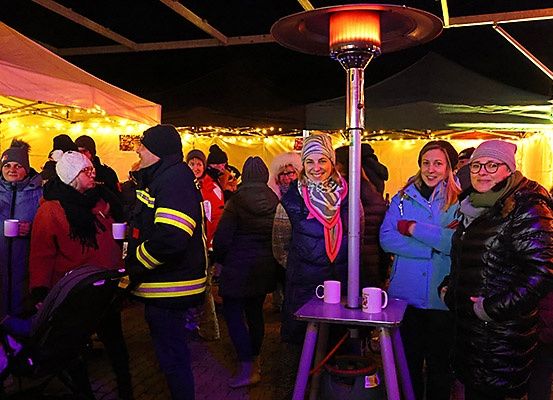 Eine Gruppe von Menschen in Winterkleidung versammelt sich bei Nacht um eine Heizung, einige mit Tassen in der Hand. Sie scheinen eine festliche Veranstaltung im Freien zu genießen.