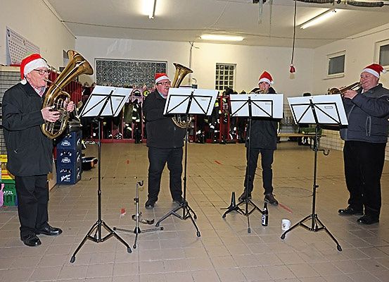 Vier Männer mit Weihnachtsmützen spielen Blechblasinstrumente in einem gefliesten Raum und stehen um Notenständer herum.