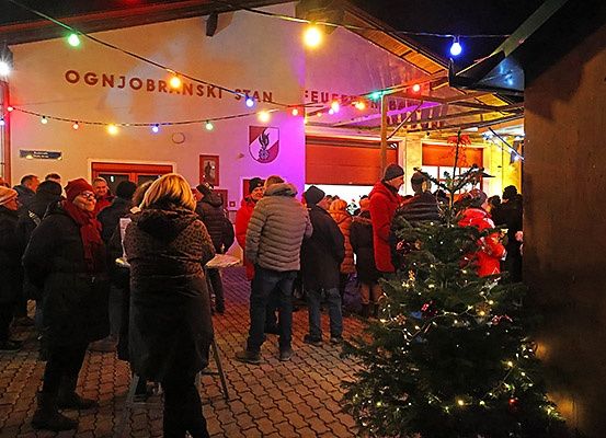 Eine Menschenmenge steht vor einem Gebäude mit festlichen Lichtern. Das Gebäude hat ein Schild, das Ognjobransk Stan steht. Ein Weihnachtsbaum ist im Vordergrund mit Lichtern geschmückt.