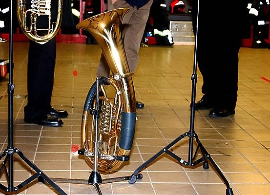 Eine goldene Tuba steht auf einem schwarzen Stativ in einem gefliesten Raum. Zwei Personen in schwarzen Hosen und Schuhen stehen dahinter. Im Hintergrund sind rote Wände und Feuerwehrausrüstung zu sehen.
