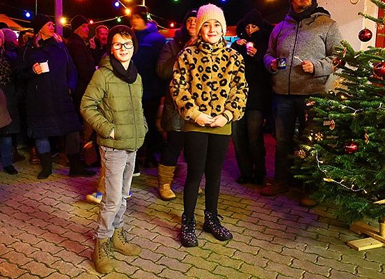 Zwei Kinder stehen vor einer Menschenmenge auf einem Weihnachtsmarkt, mit einem geschmückten Weihnachtsbaum in der Nähe. Die Menschen im Hintergrund tragen Winterkleidung.
