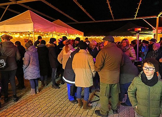 Eine Menschenmenge, wahrscheinlich auf einem Weihnachtsmarkt, steht unter beleuchteten Zelten. Einige tragen Brillen, Mützen und haben Taschen bei sich. Ein Kind in einer grünen Jacke sticht rechts hervor.