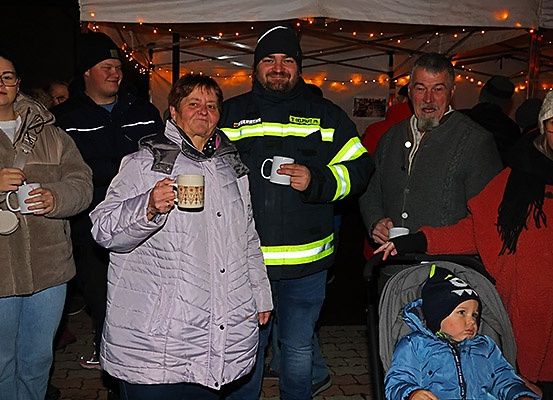 Eine Gruppe von Menschen steht unter einem Zelt. Eine Frau in der Mitte hält eine Tasse. Einige tragen Winterkleidung und Mützen. Ein Mann neben ihr trägt eine reflektierende Jacke. Ein Kind sitzt in einem Kinderwagen.