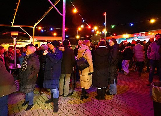 Viele Menschen sind nachts in einem Außenbereich versammelt und tragen Winterkleidung. Bunte Lichter hängen über ihnen. Einige Leute gehen, und einige stehen in der Nähe des Mülleimers.