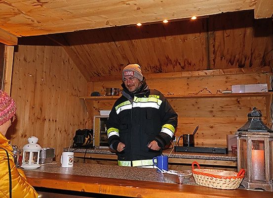 Ein Mann in einer reflektierenden Jacke steht in einer Holzhütte mit Theke, Lampe und Korb. Eine weitere Person mit Mütze sitzt links.