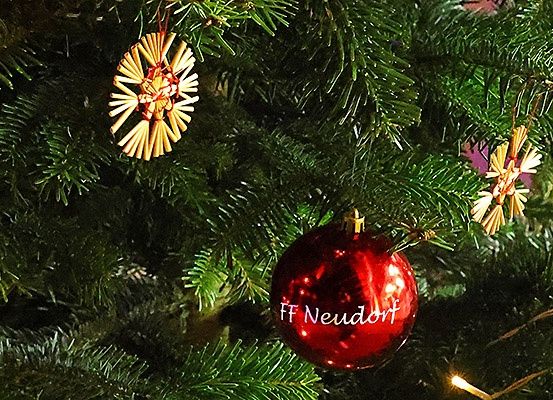 Eine rote Weihnachtskugel mit 'FF Neudorf' steht an einem grünen Weihnachtsbaum. Andere Ornamente sind ebenfalls sichtbar.