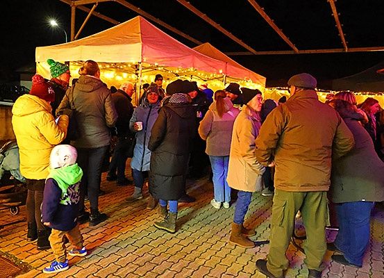 Eine Gruppe von Menschen versammelt sich auf einem Nachtmarkt unter einem Baldachin, einige tragen Winterkleidung und Mützen. Ein Kinderwagen ist in der Nähe, und der Bereich ist von dekorativen Lichtern beleuchtet.