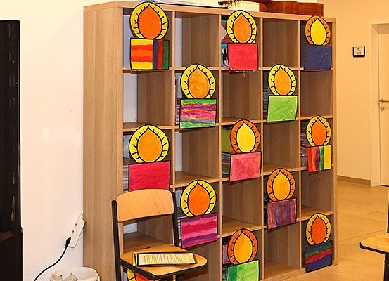 Ein hölzerne Bücherregal mit mehrfarbigen Lampendesigns. Ein Stuhl mit einem Papier darauf. Ein Lautsprecher ist an der Wand montiert.