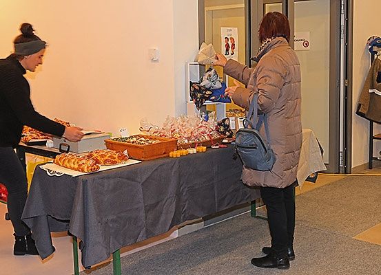 Zwei Frauen stehen bei einem Tisch mit verschiedenen Gegenständen darauf, einschließlich Brot, Süßigkeiten und einem Schild. Eine Frau spricht mit der anderen, die die Gegenstände betrachtet.