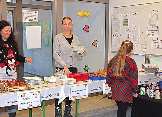 Drei Frauen stehen an einem Tisch mit Essen und Getränken. Eine Frau hält einen Stapel Teller in der Hand, während eine andere Frau sie ansieht. Der Tisch hat ein Schild mit der Aufschrift Kaffee und Mehlspeise. Dahinter befindet sich eine weiße Tafel mit angehefteten Papieren.