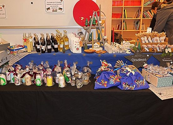 Ein Tisch mit blauer Tischdecke ist mit verschiedenen Geschenkartikeln und Behältern bedeckt. Flaschen, kleine Tüten und Geschenkboxen sind in Reihen angeordnet. Hinter dem Tisch befindet sich eine weiße Wand mit Text und einer runden Dekoration.